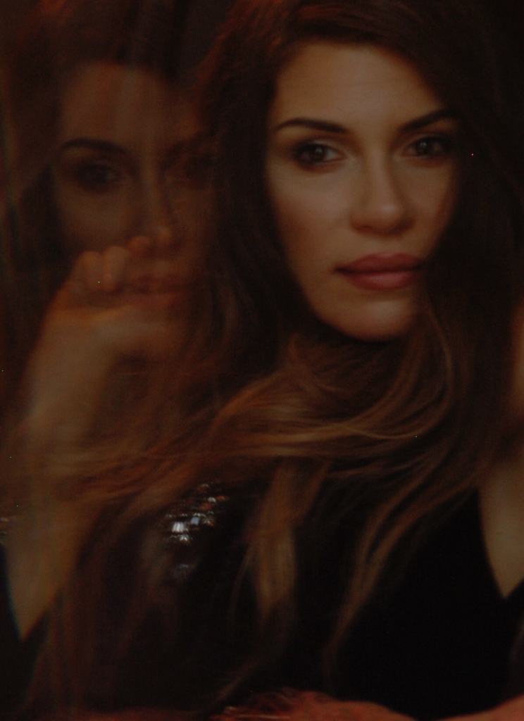 Reflection of a woman’s face in glass beside her own, creating a layered double image that evokes introspection, identity, and the fractured self.
